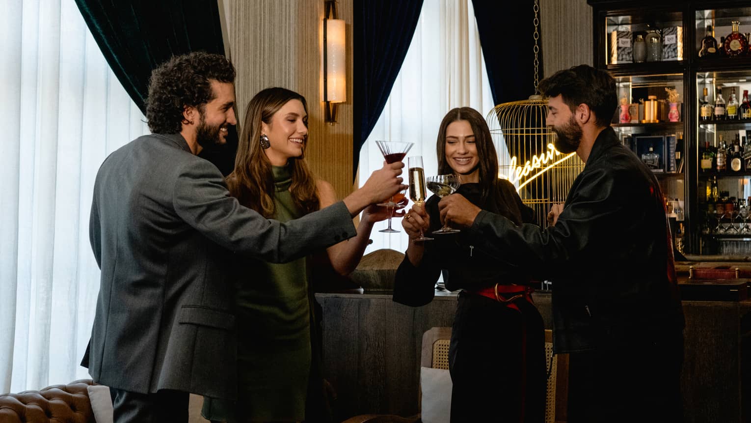 Four people raise their glasses in a toast in a cozy, softly lit lounge setting.