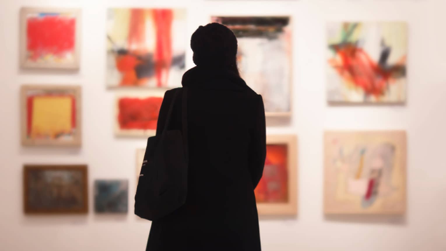 Silhouette of back of woman with hat bag looking at gallery wall with colourful art