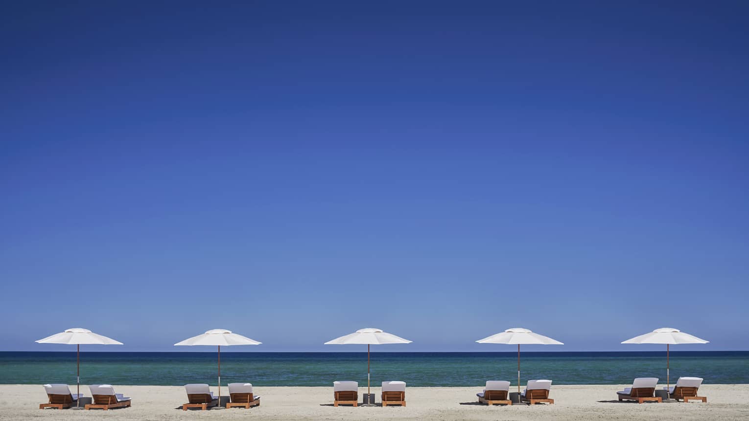 Row of white patio umbrellas, chairs on beach against striking blue sky