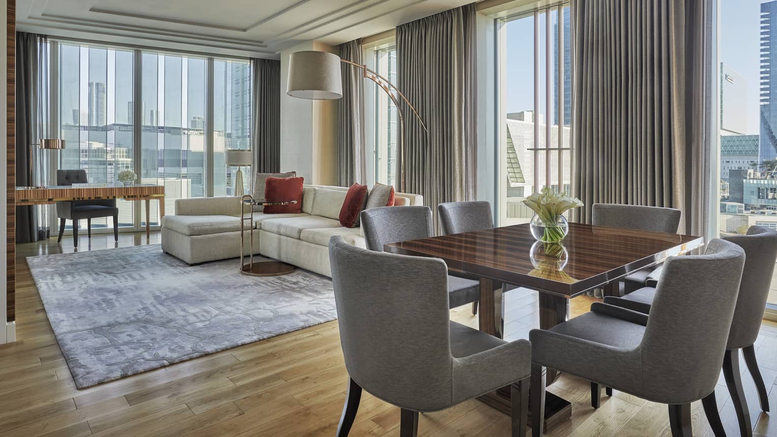 Spacious modern living and dining area with a cream sectional sofa, red cushions, a wooden dining table and floor-to-ceiling windows offering a city view