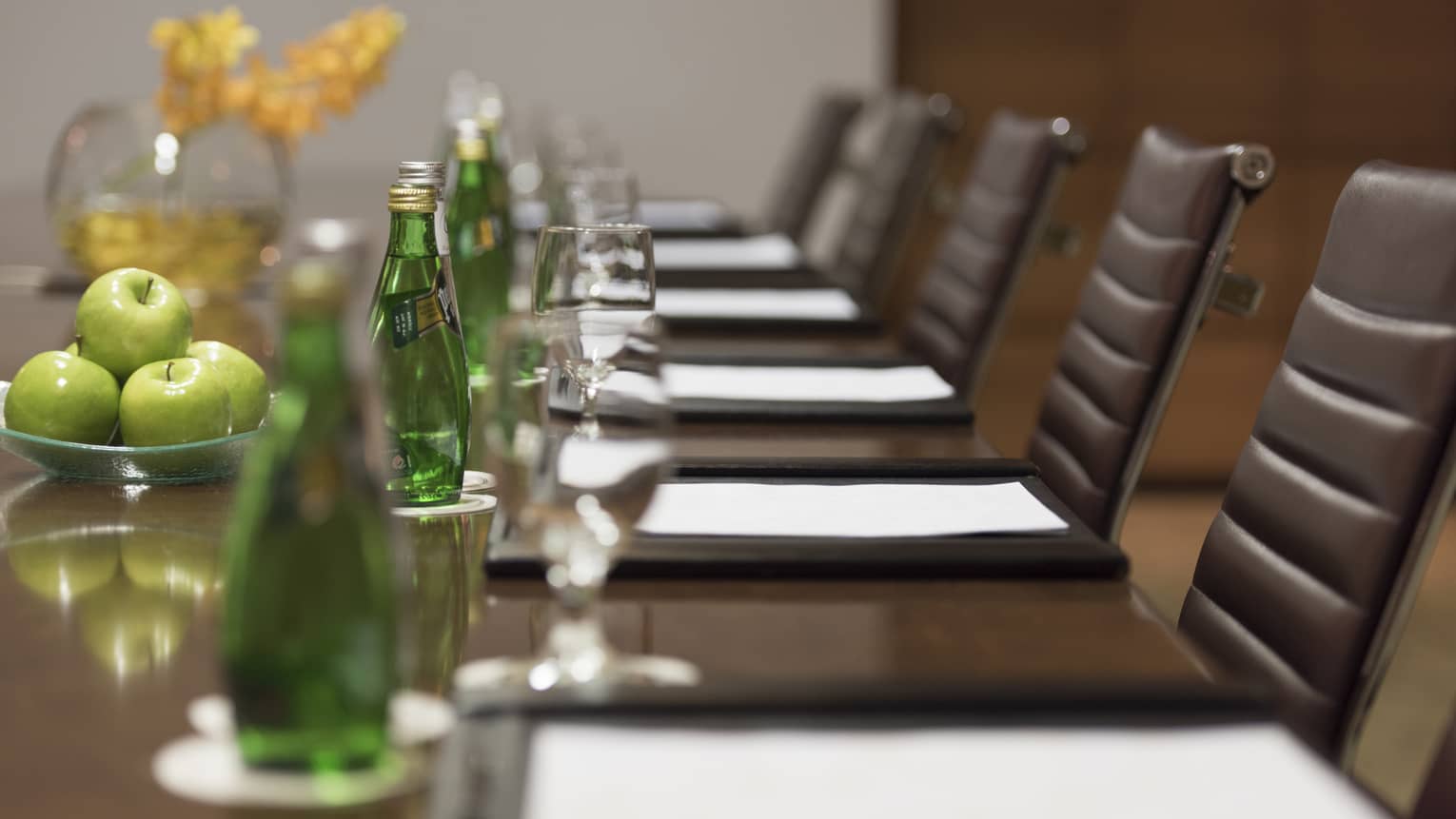 Close-up of business meeting table row of chairs, paper agendas, green glass bottles, apples