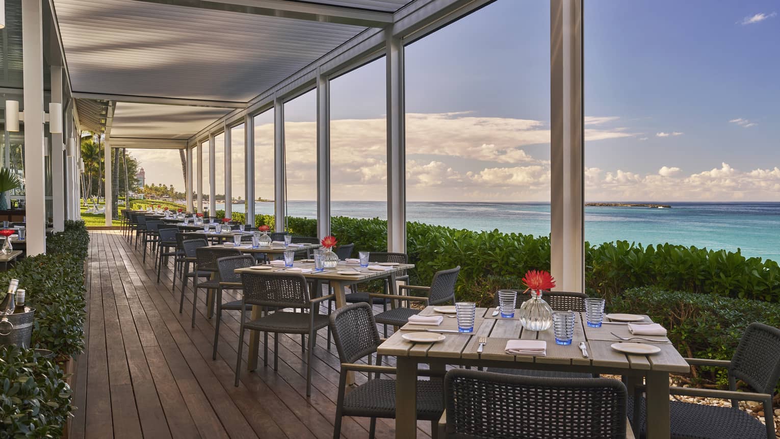 An outdoor eating area with square wood tables next to plants and the ocean.