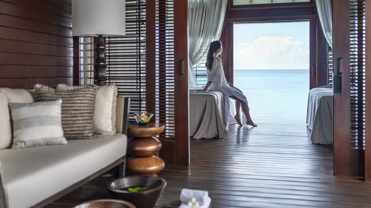 A woman leans on a massage bed on the far side of a spa treatment room and looks out through a large doorway onto the vast blue ocean