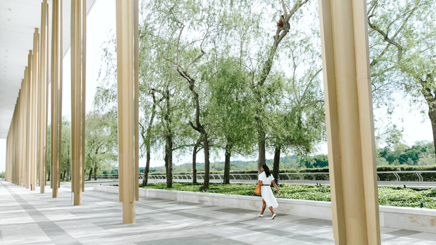 A woman inn a white dress holding an orange bag walking along beige columns outside near trees.