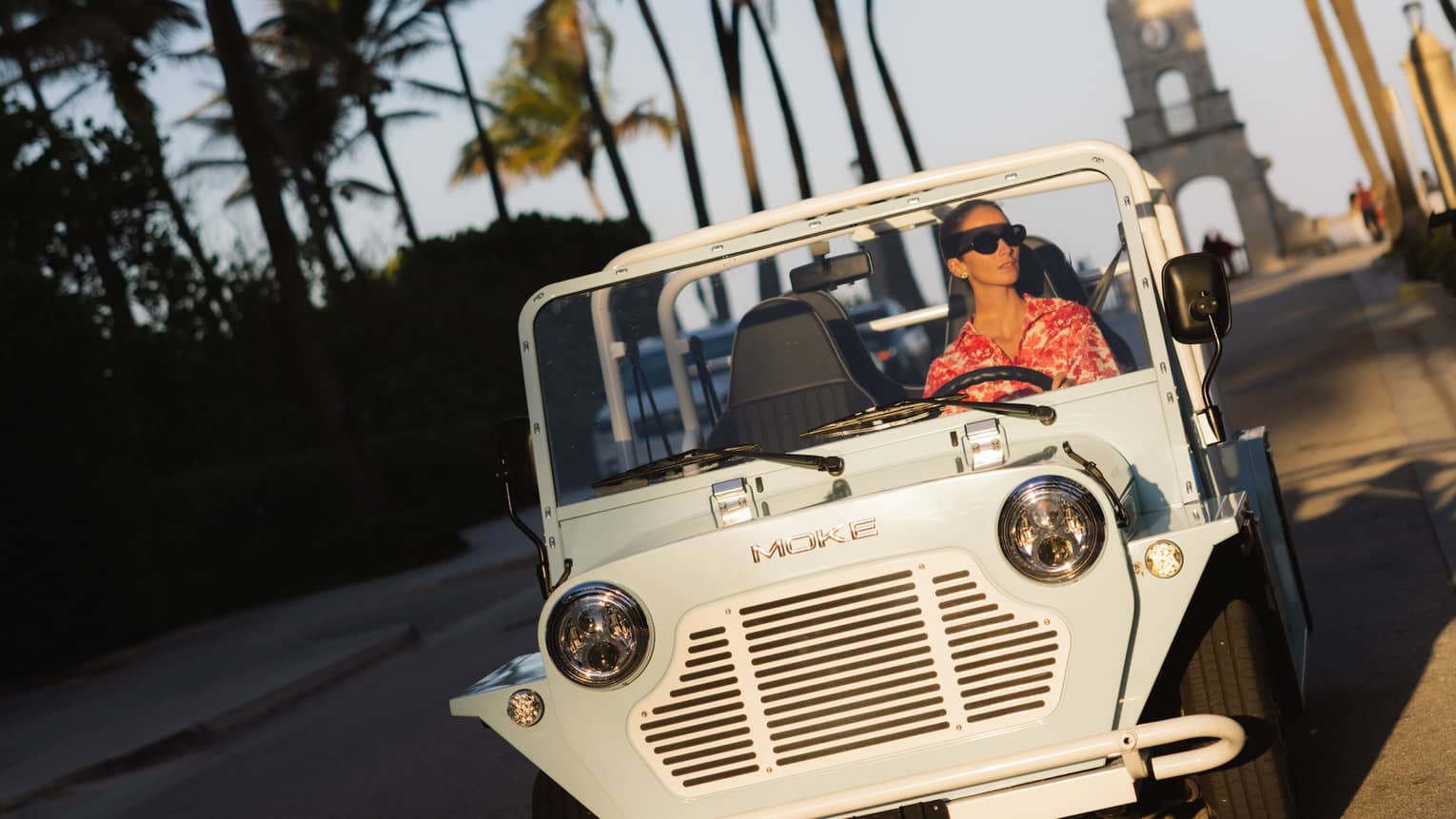 Guest wearing a red-and-white tropical shirt rides in a white MOKE along a palm tree?lined street at dusk