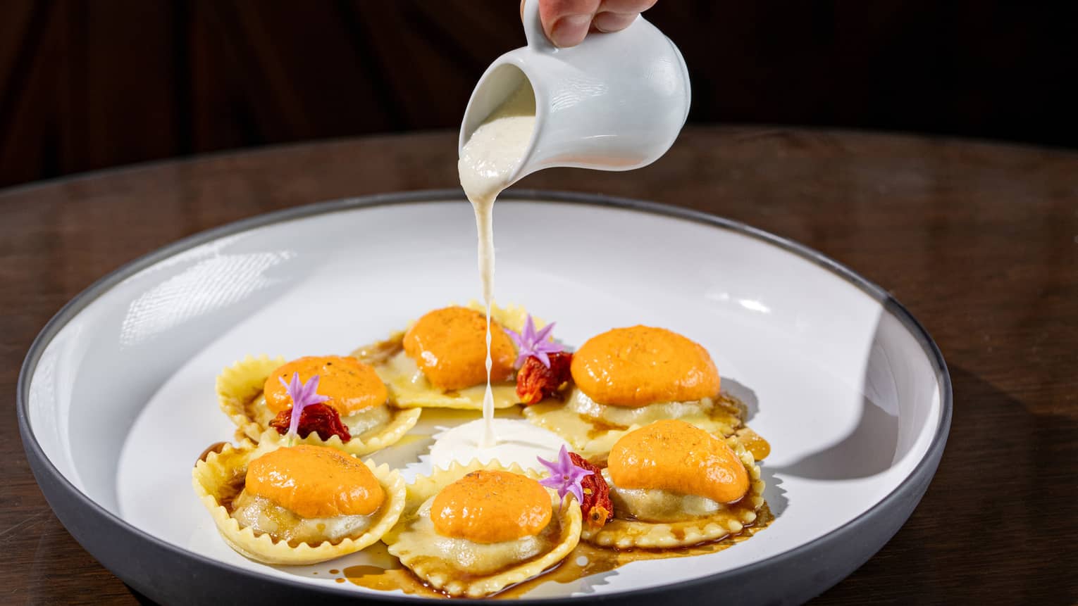 Six ravioli in a circle, topped with tomato espuma, and a hand pouring a white sauce over