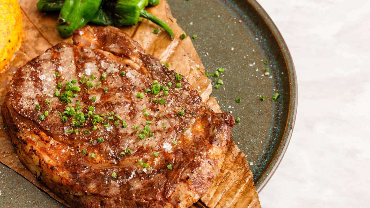 Rib-eye steak served on a dark plate with grilled green peppers and a small side bowl of red sauce