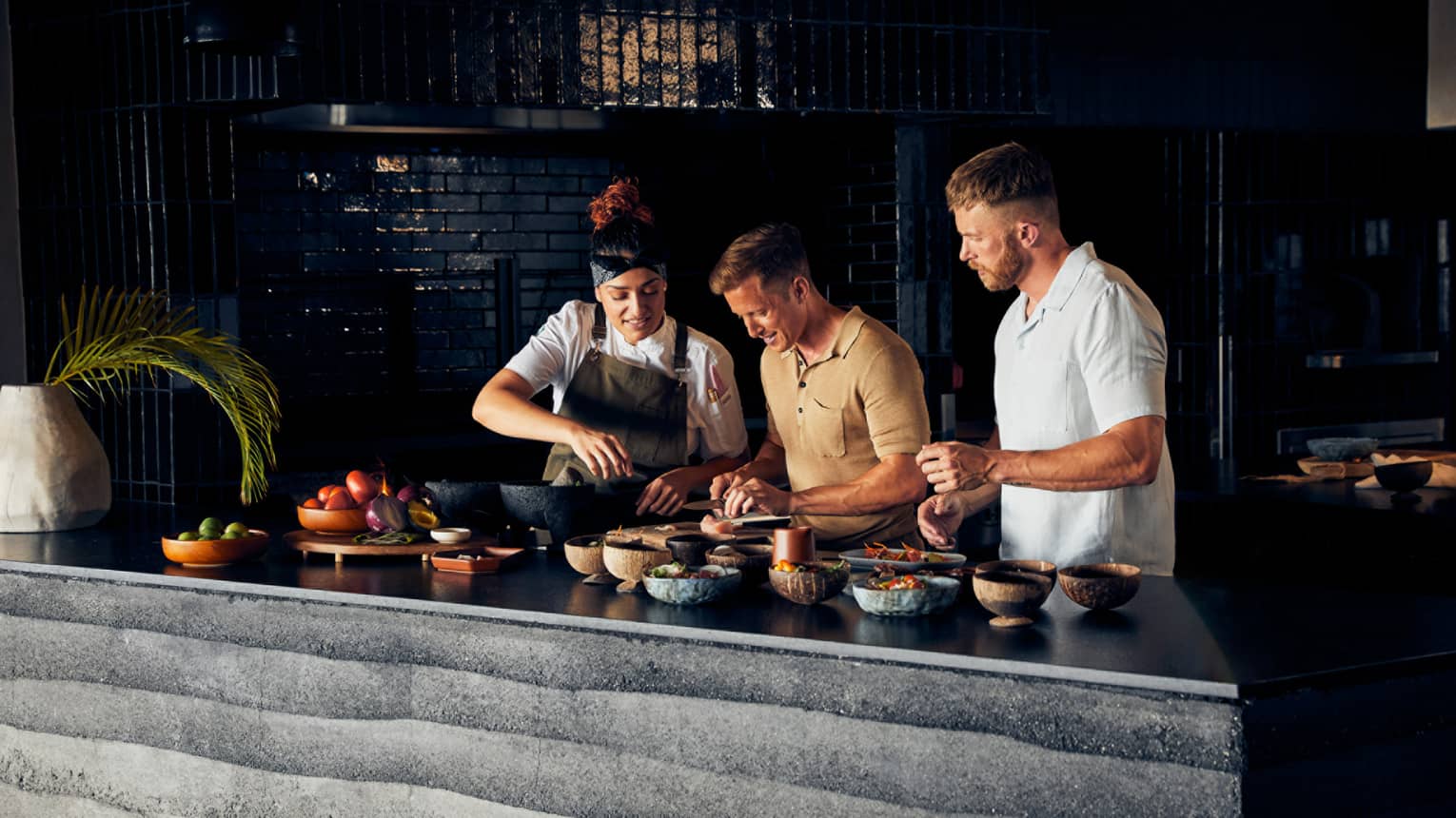 Two men cooking with a chef in a dimly lit room.