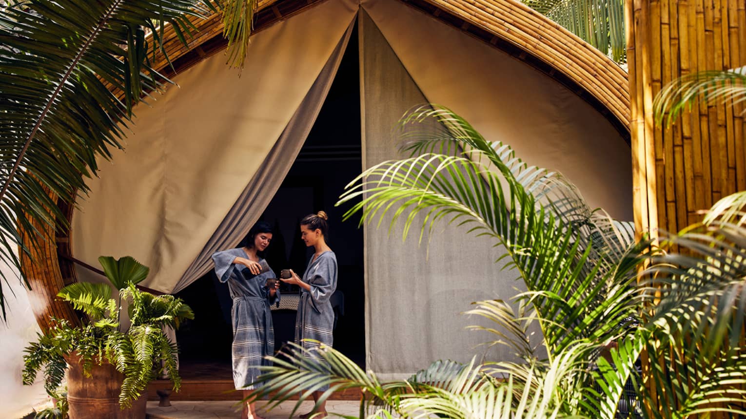 Two women in spa robes stand outside a luxury spa tent, one pouring tea for the other, surrounded by lush greenery in a peaceful setting.