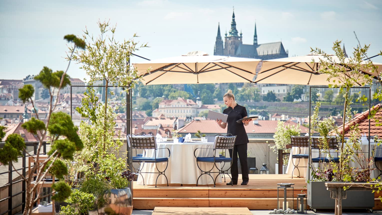 Hotel staff sets dining table under umbrella on rooftop patio overlooking sunny Prague city 