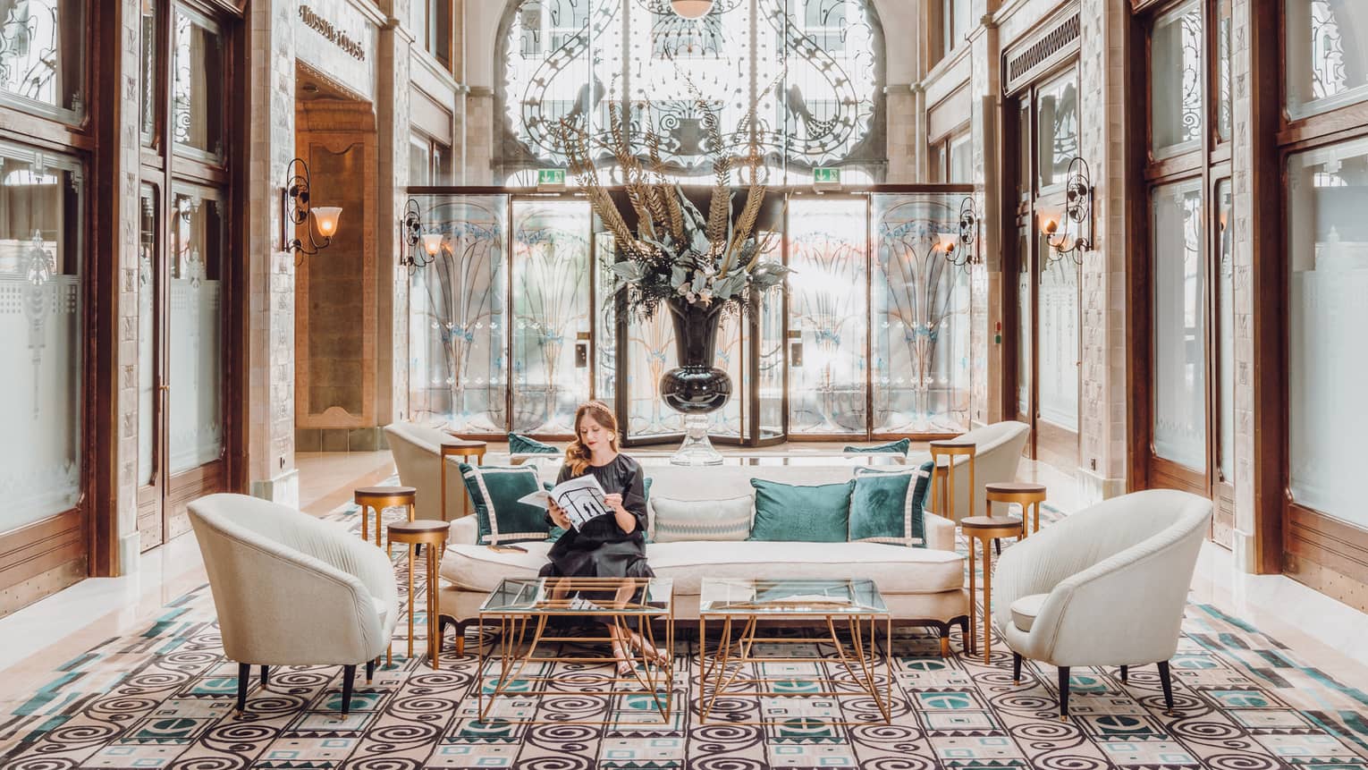 Elegant hotel lobby with arched glass ceiling, decorative floor tiles, plush seating and a large floral arrangement. A person sits on a sofa reading a magazine.