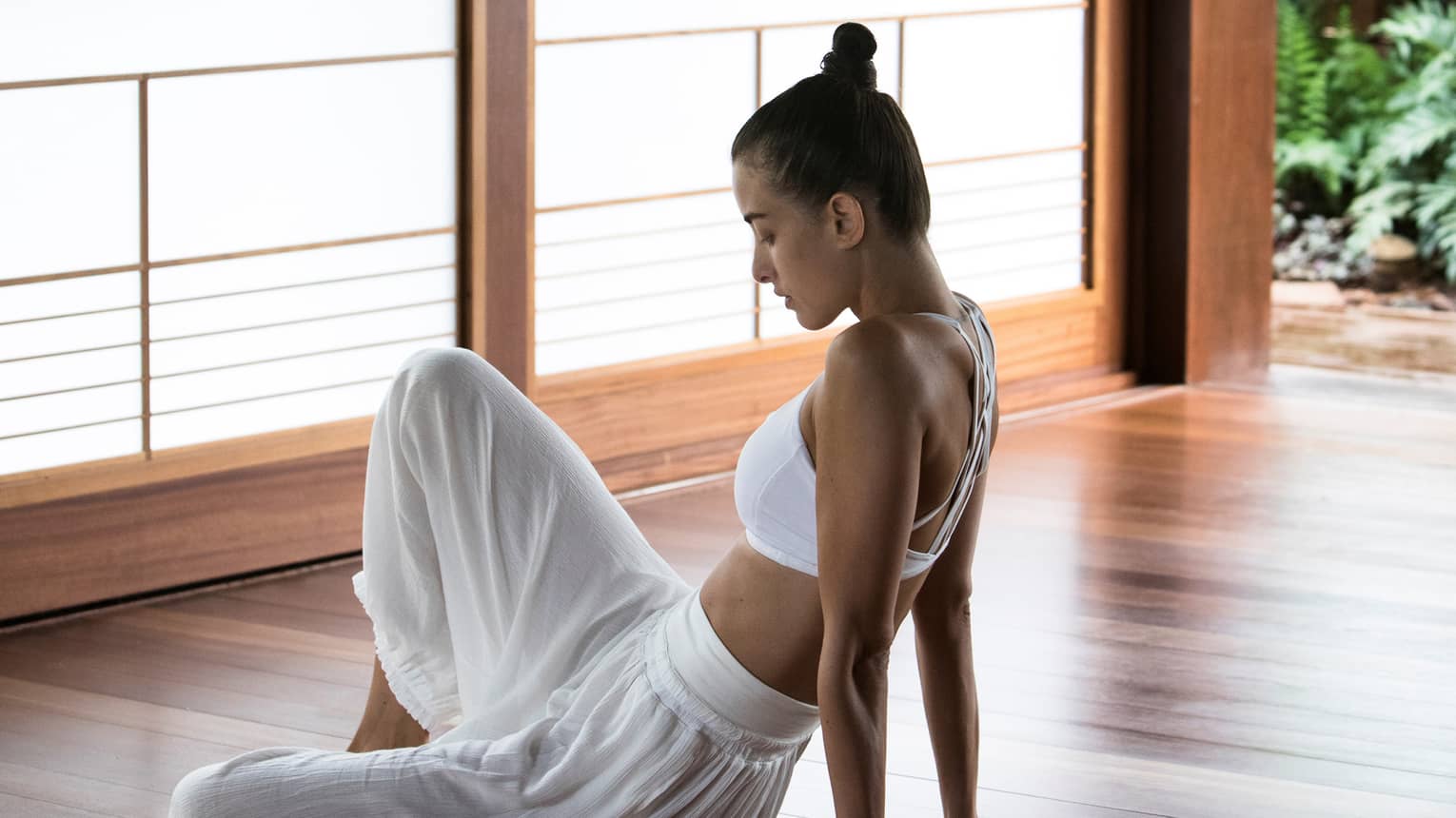 A woman dressed in workout clothes sits on the floor of a room with wooden walls and windows overlooking lush greenery