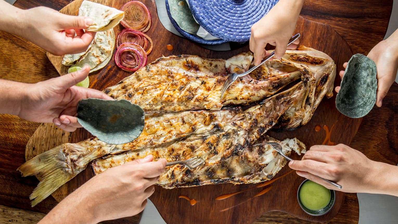 Aerial view of table, hands holding forks, digging into whole grilled fish on platter