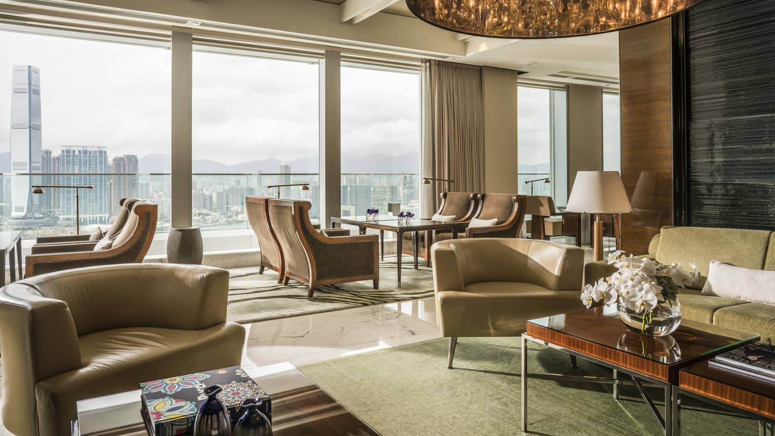 A spacious, modern lounge area with large windows offering a panoramic cityscape view. The room features comfortable armchairs, sofas, wooden furniture.