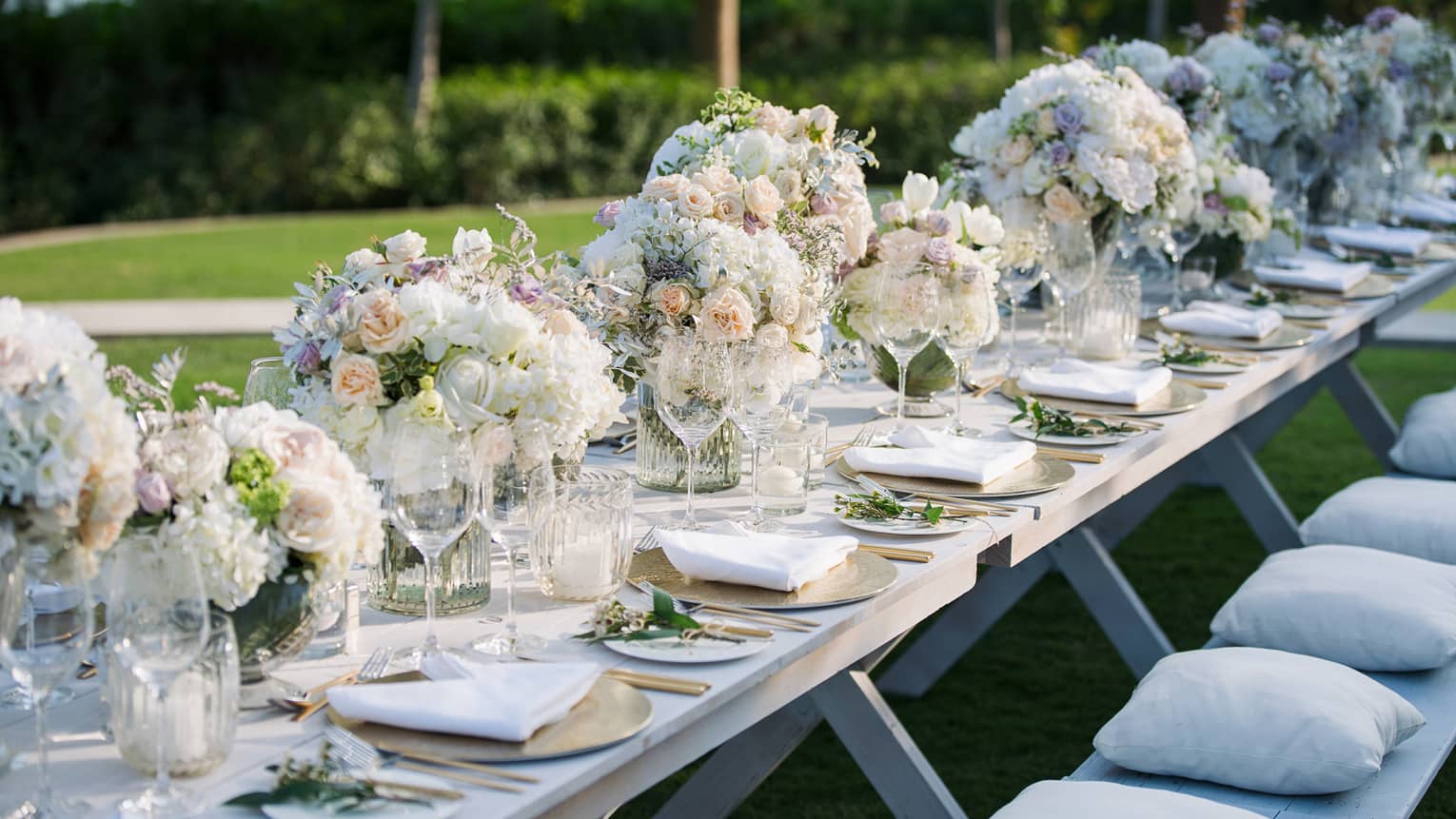 A banquet style table is set with gold chargers, white plates, tableware and large white bouqets