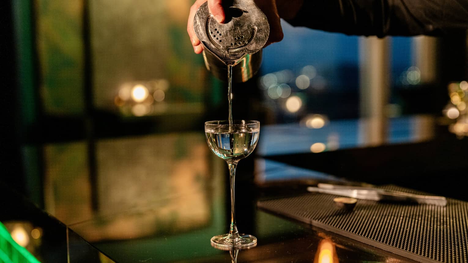 Bartender's hand pours liquid from a silver shaker into a tall stemmed cocktail glass