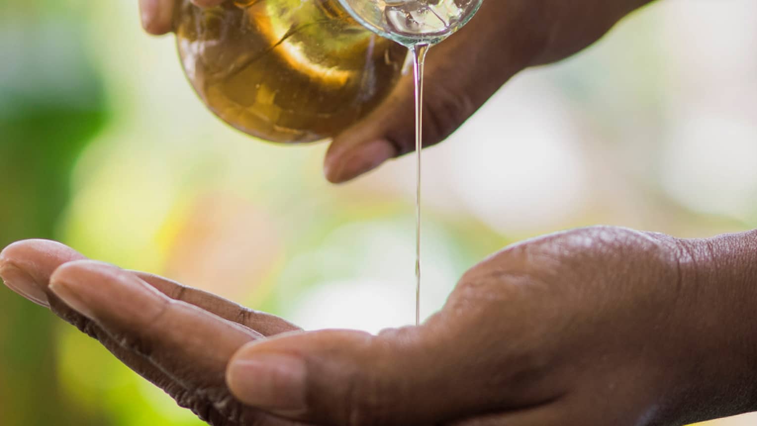 Golden oil flows from a small carafe held in one hand onto the other, a small wooden bowl underneath to catch the overflow.