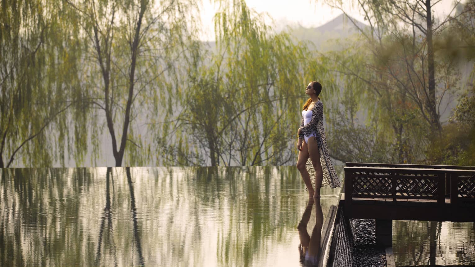 Woman wearing swimsuit, sheer cover walks by infinity swimming pool, water reflecting trees