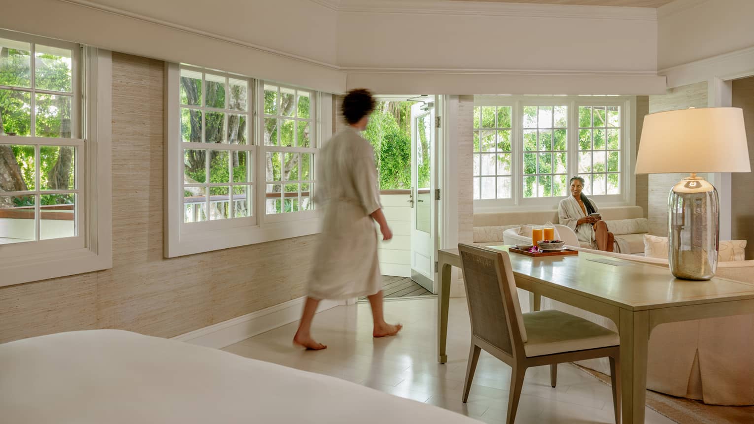 A man wearing bathrobe walks in a hotel suite that features a desk, living room area and opens to gardens