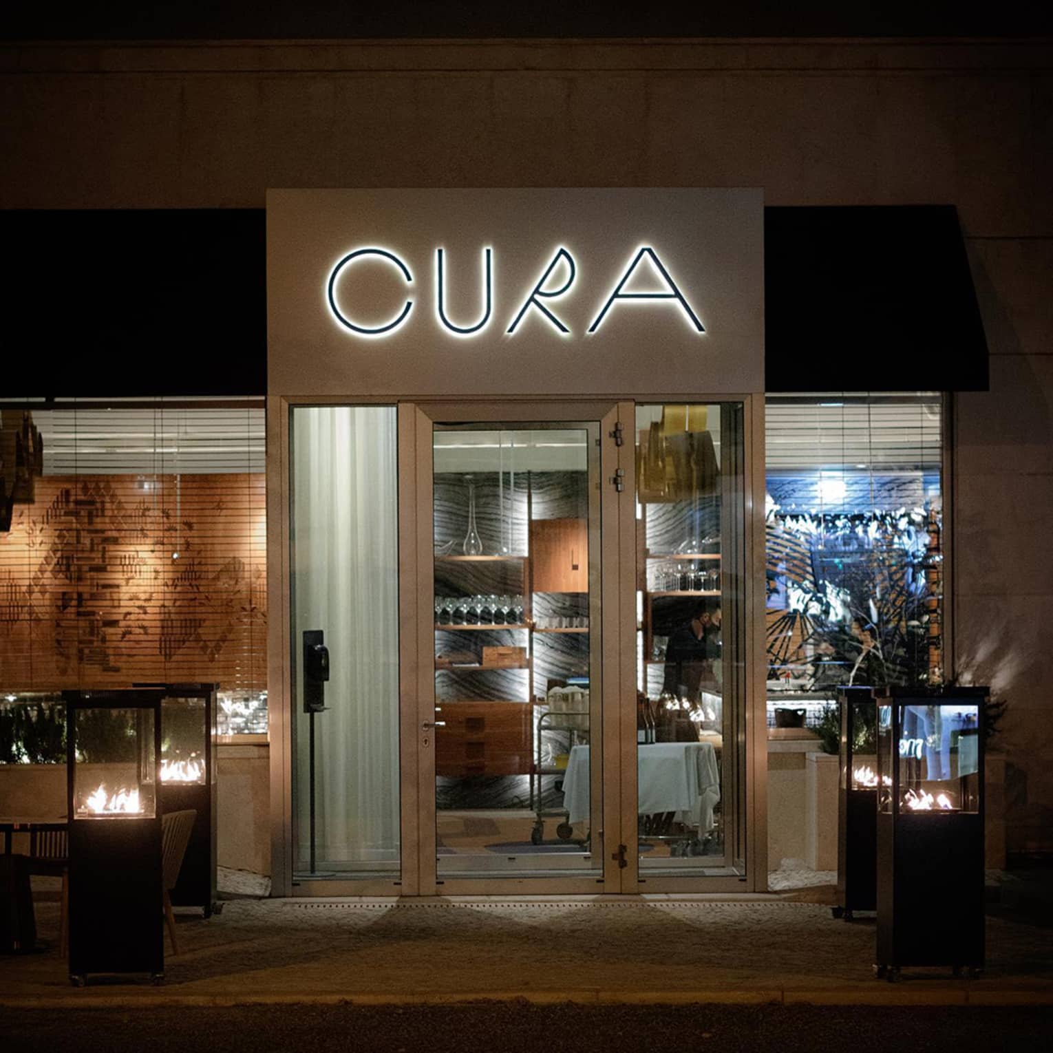 Nighttime exterior of CURA restaurant, featuring modern glass doors, a minimalist neon sign and warm fire-lit outdoor lanterns, creating an inviting atmosphere.