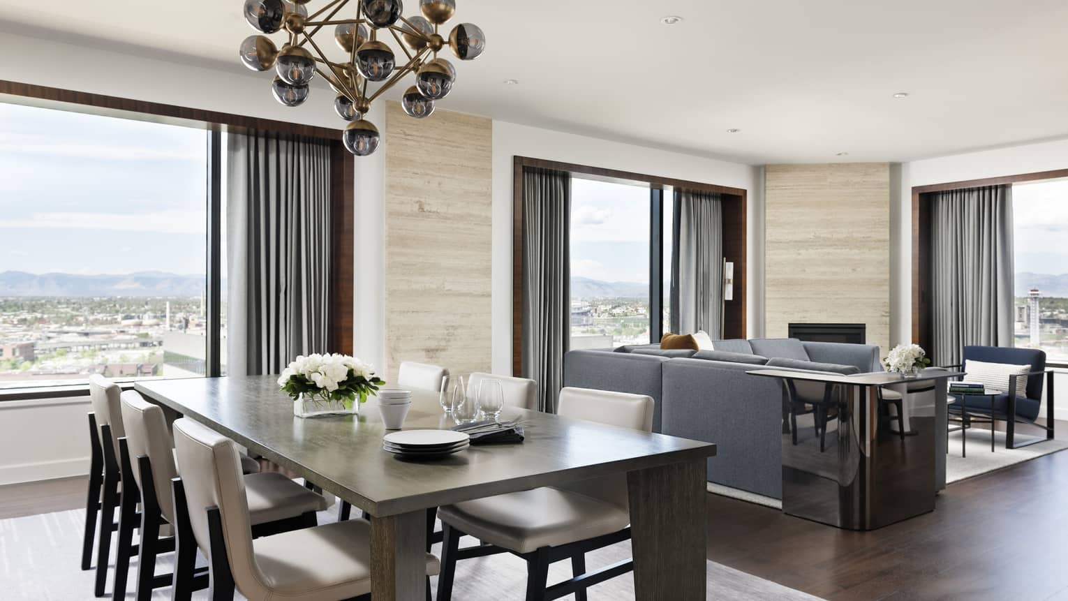 A large rectangular dining room table with a gold chandelier next to large windows and a seating area with sofa and chairs.