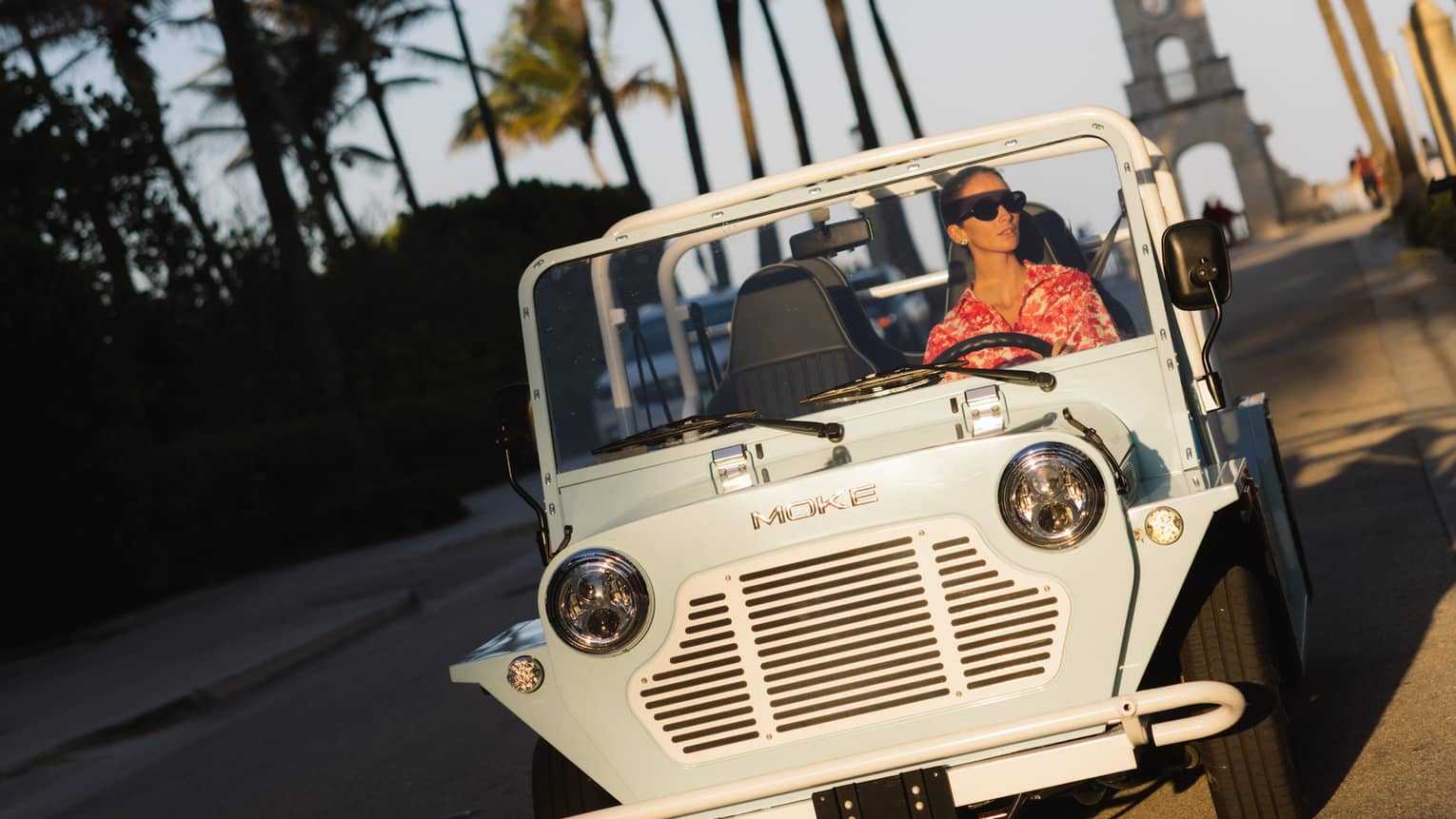 Guest wearing a red-and-white tropical shirt rides in a white MOKE along a palm tree?lined street at dusk