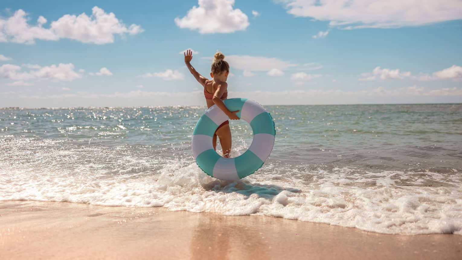 Young child walks into the ocean on a beach holding a turquoise and white striped floatie