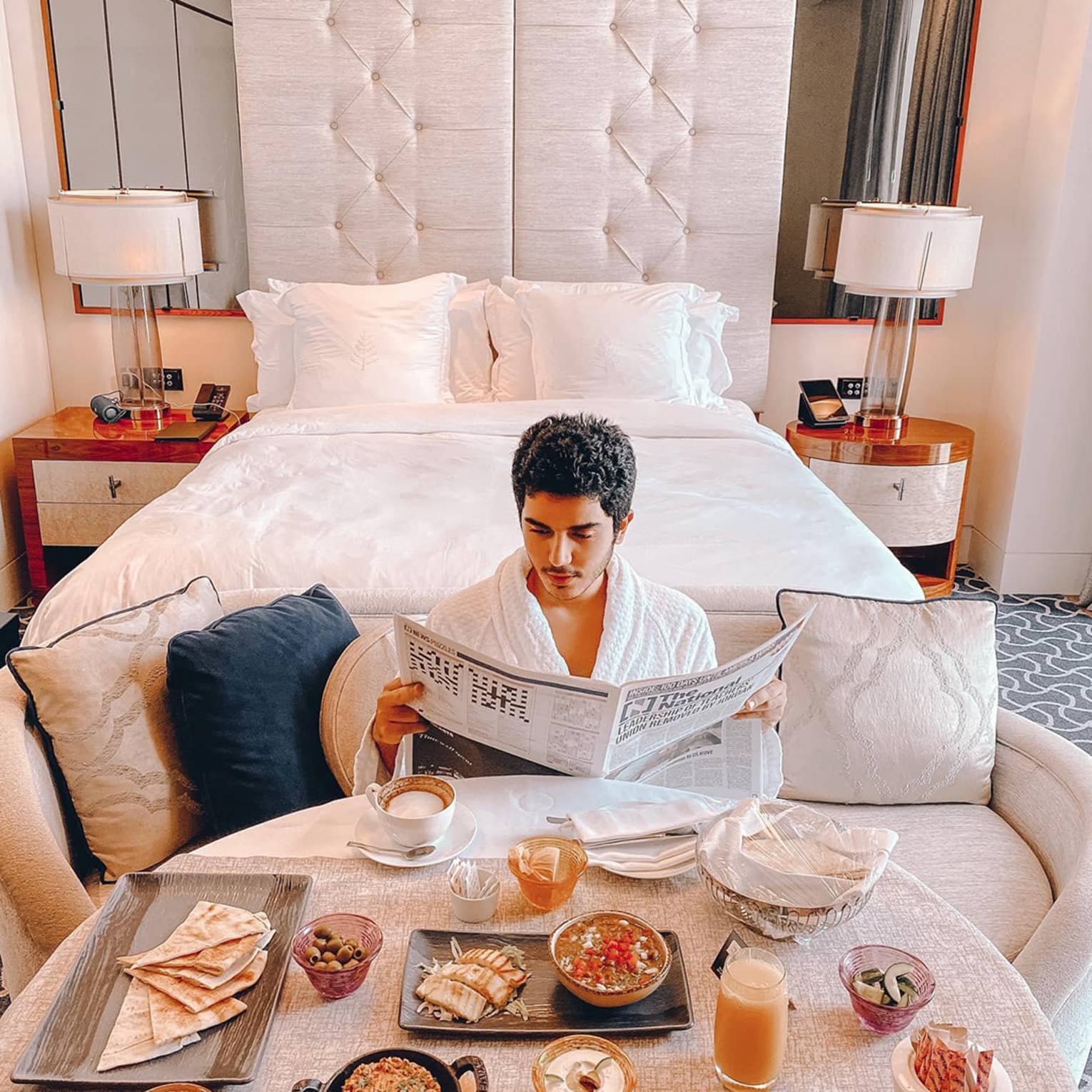 Man wearing bath robe reads newspaper in front of breakfast table, hotel bed