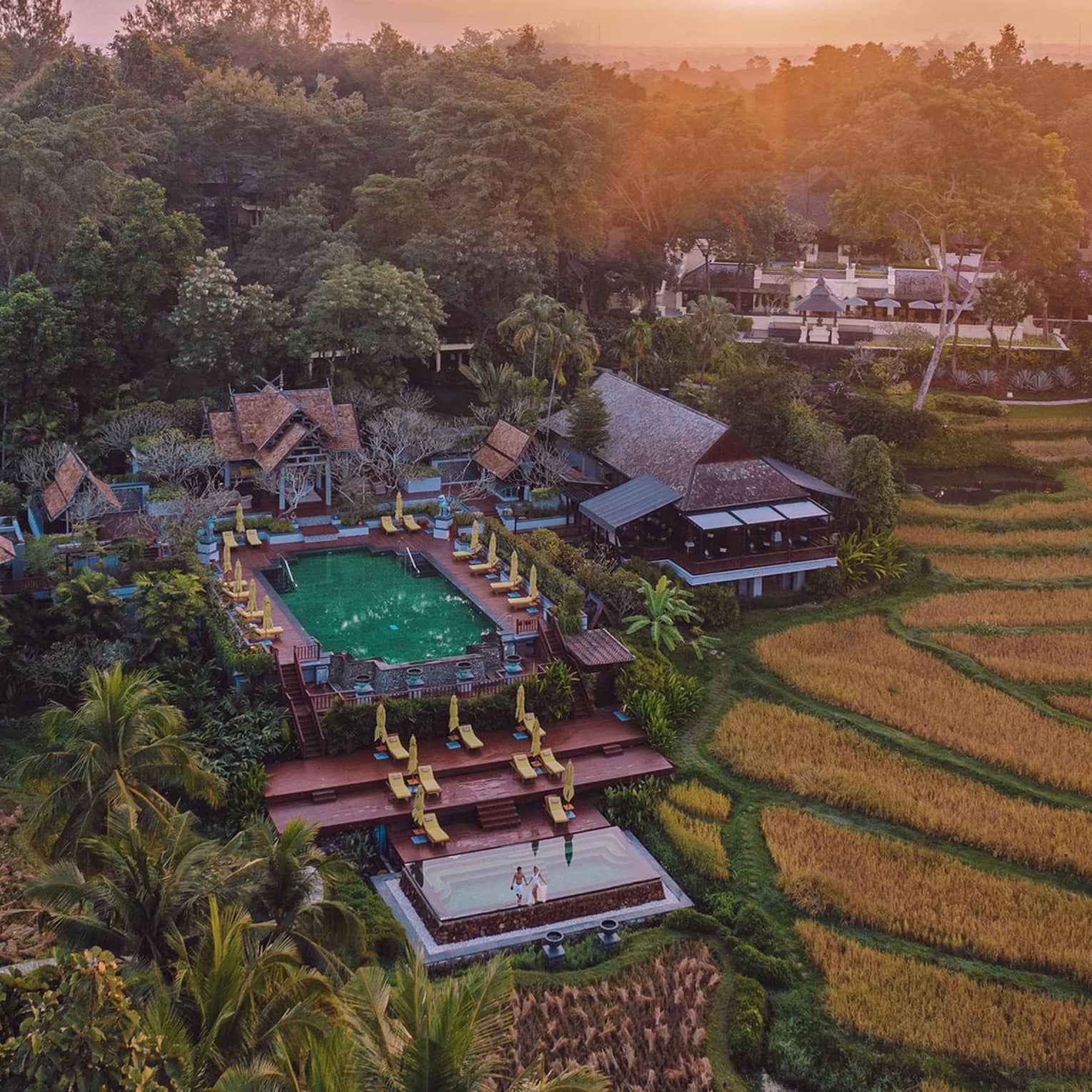 Aerial view of a resort with terraced pools, sun loungers and traditional architecture surrounded by rice fields at sunset.