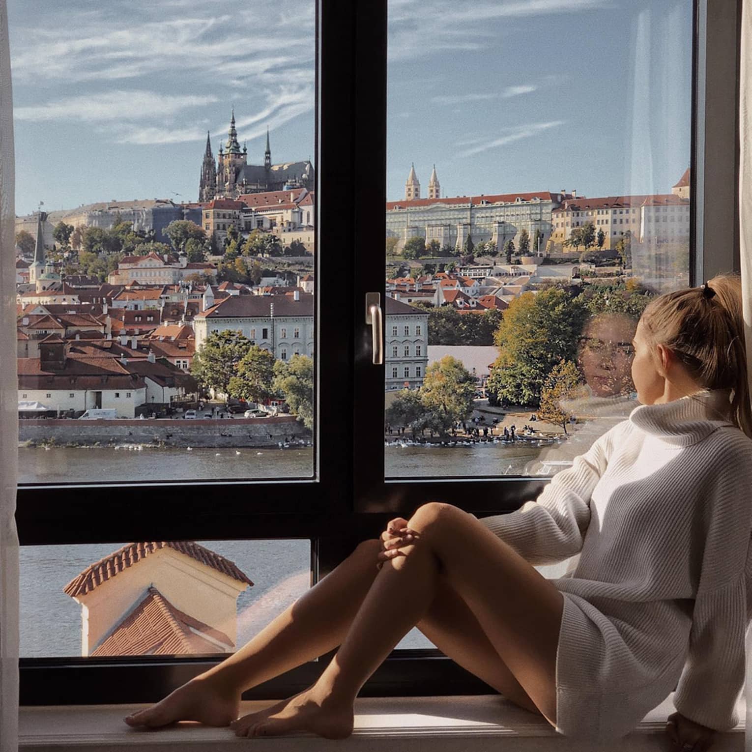 A person sitting by a window, looking out at a scenic view of historic buildings and a river.