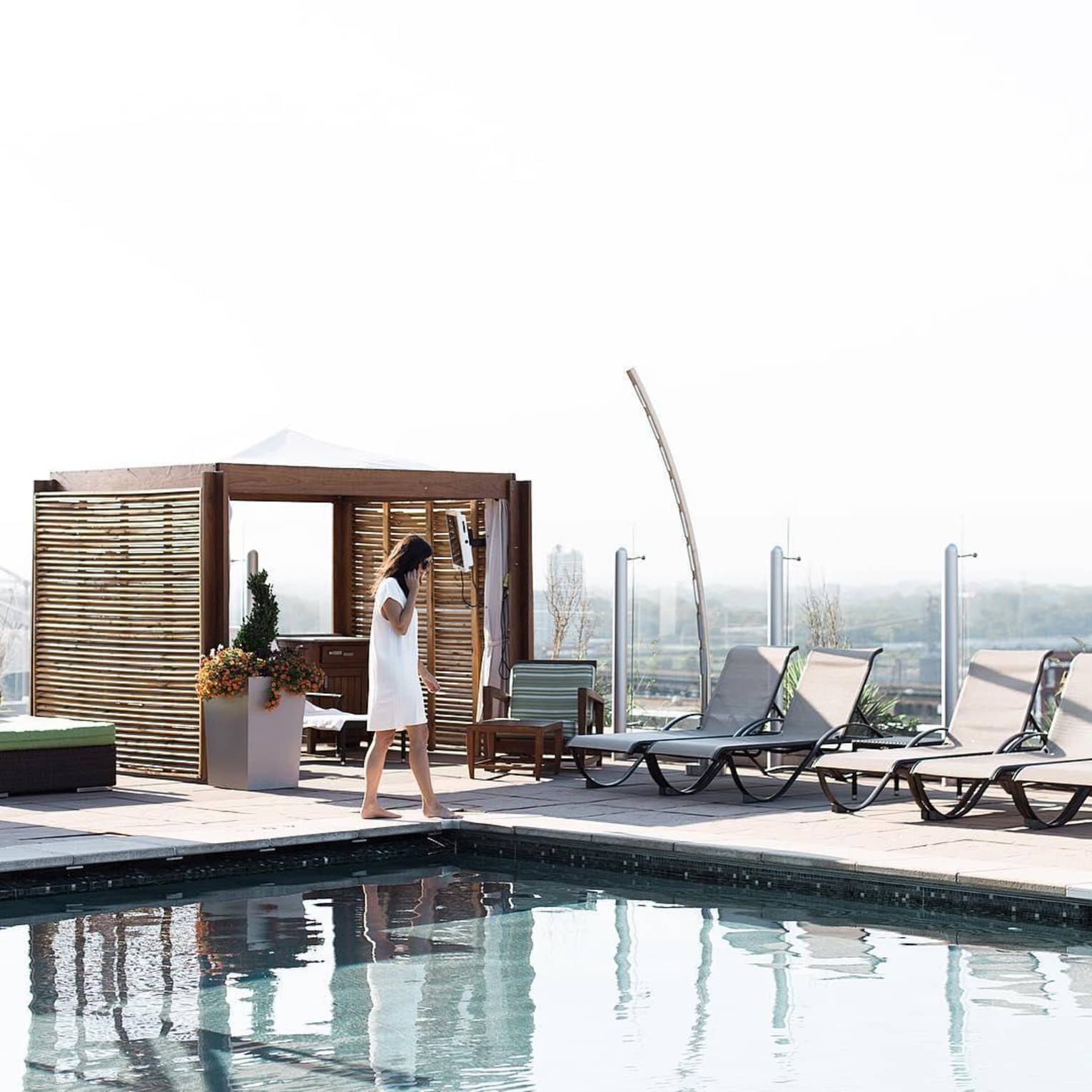 Person in white dress walking barefoot by a hotel rooftop pool with lounge chairs and a private cabana in the background