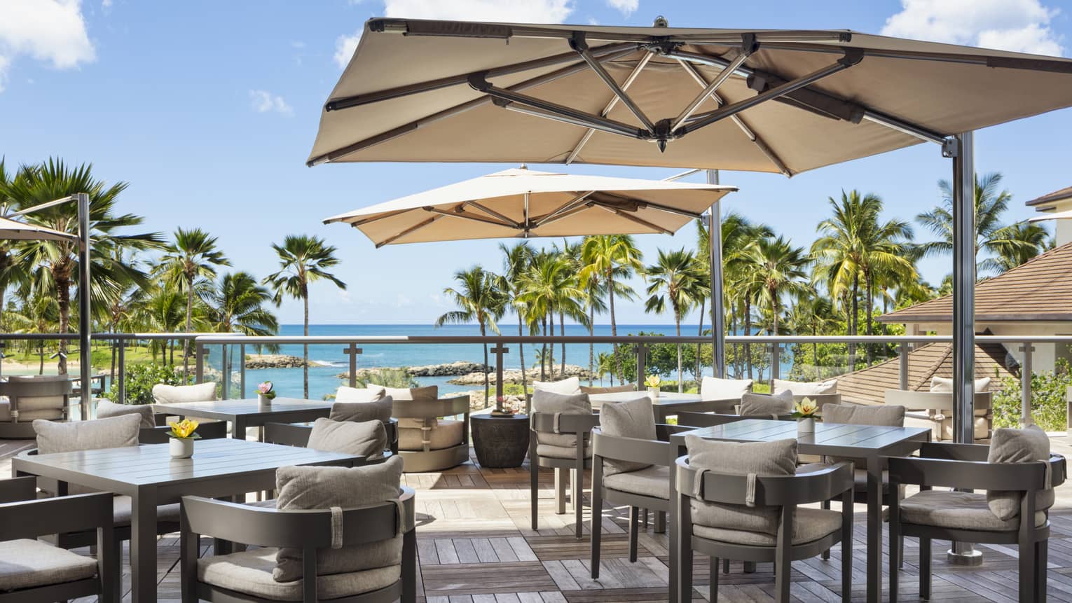 An outdoor dining space has wood deck floors, square tables shaded by large sun umbrellas and chairs with beige cushions and offers a view of palm trees and the ocean.