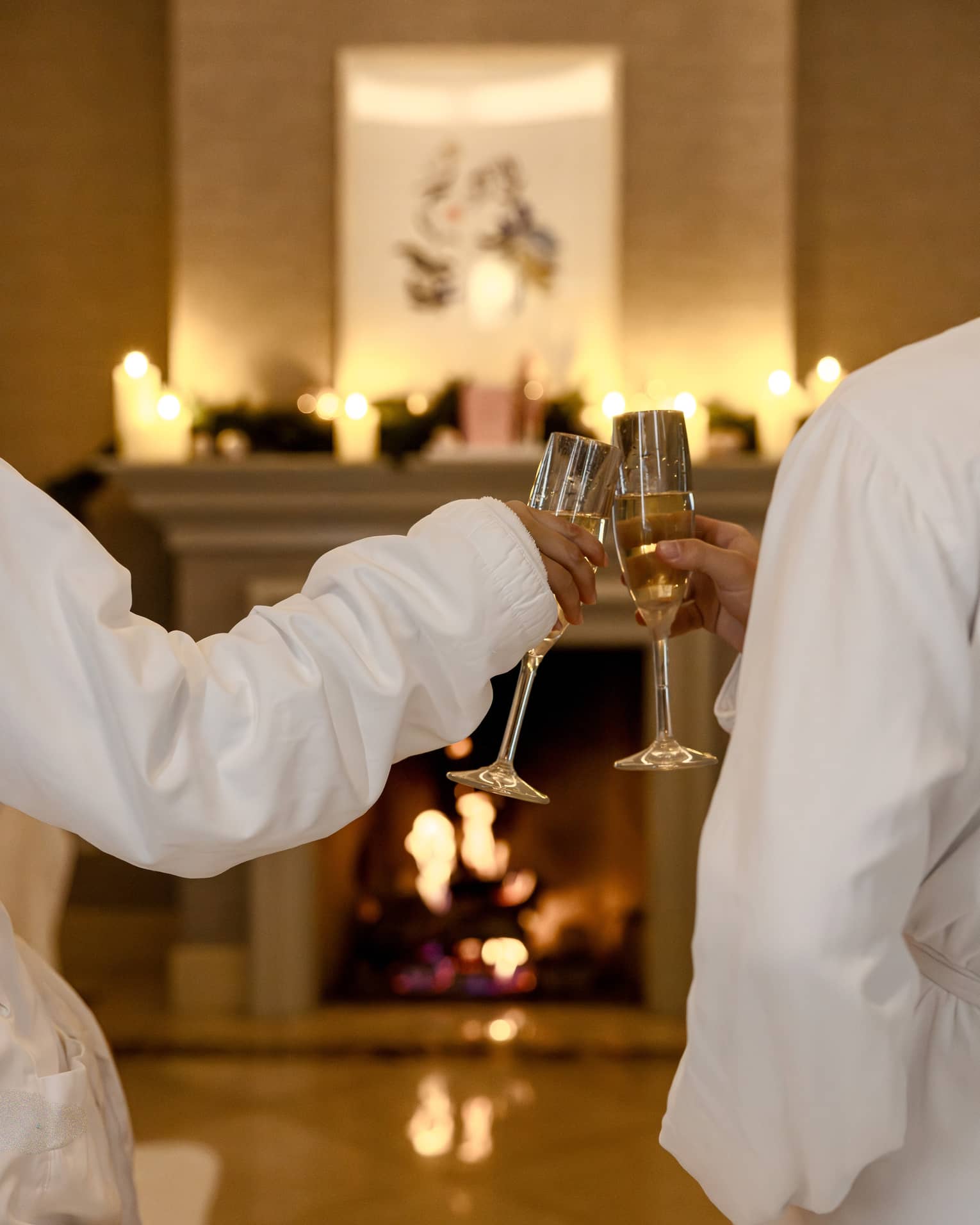Two guests wearing white robes sit in front of a fireplace toasting each other with glasses of Champagne