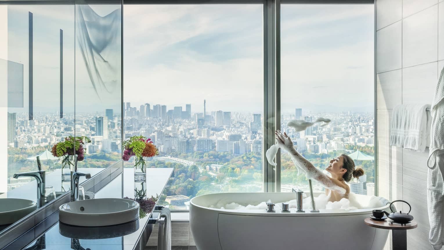 Woman in soaking tub in guest bathroom, floor-to-ceiling windows with city views