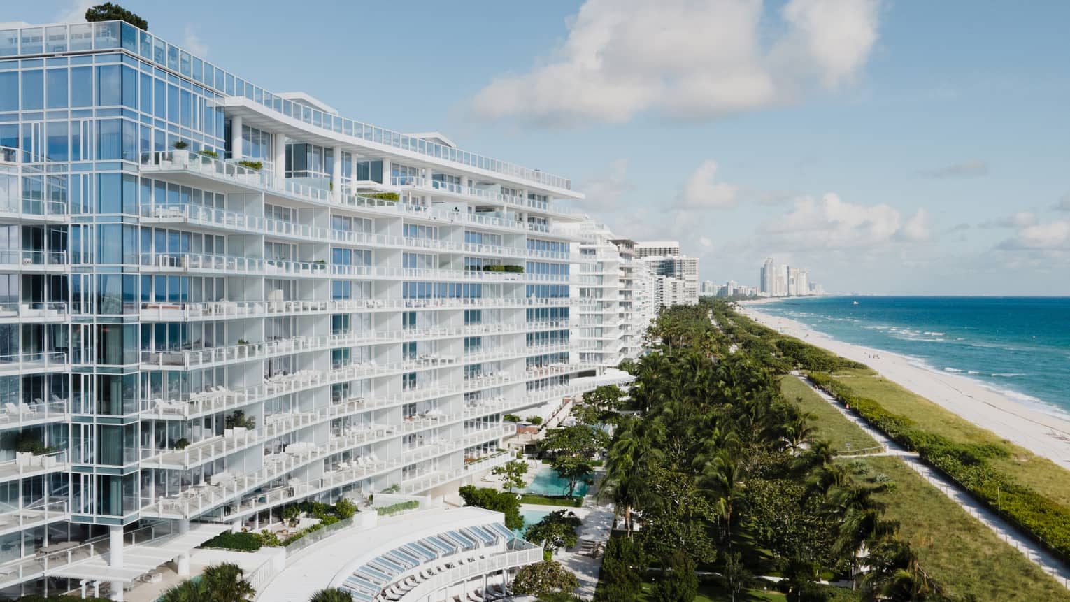 Luxury beachfront hotel with modern glass architecture, lush greenery, a pool area and a view of the ocean and sandy beach under a blue sky