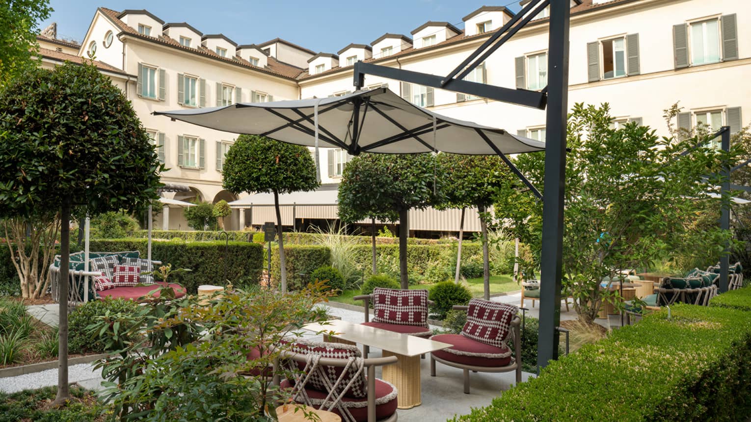 Elegant courtyard vignette under tan umbrella surrounded by hedge and topiaries with hotel in backdrop