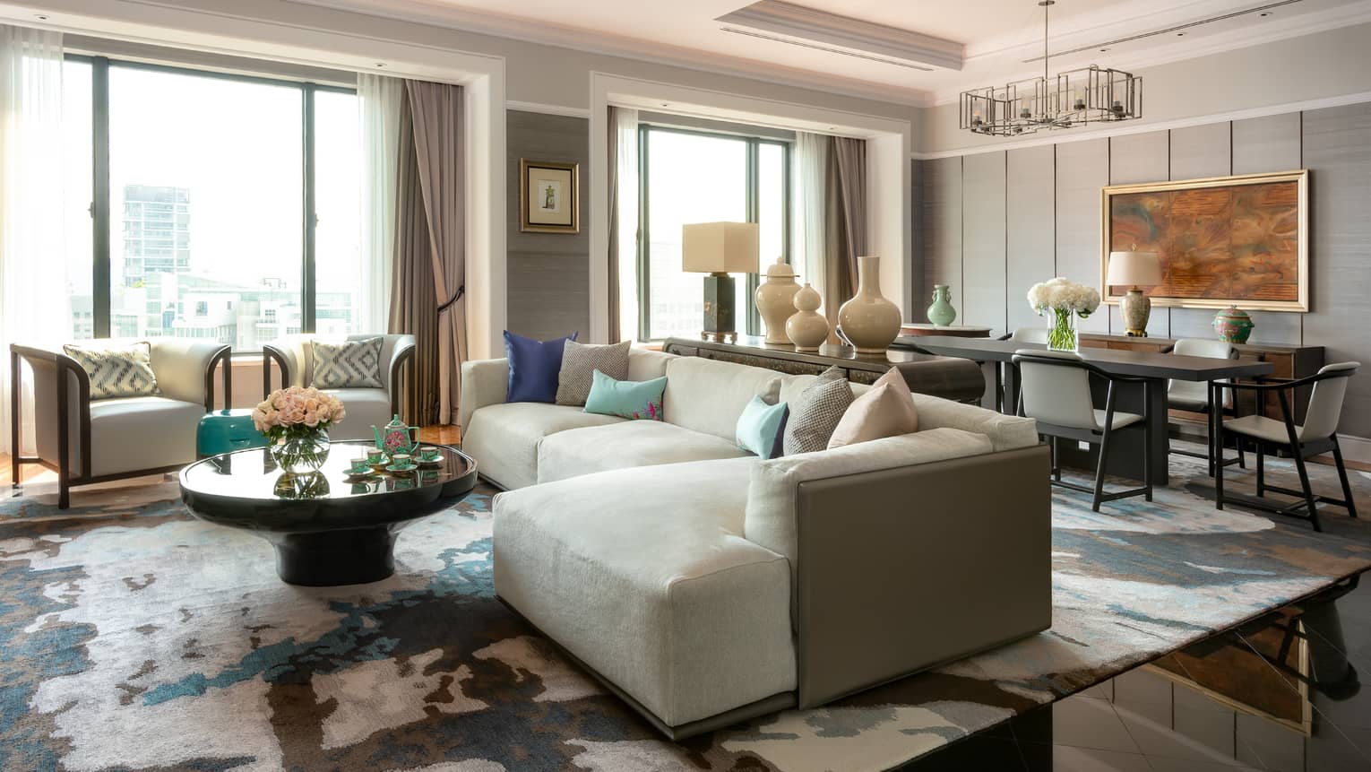 A spacious, luxurious living room with large windows, light grey sectional sofa, round coffee table and modern décor, including vases and artwork on the walls.