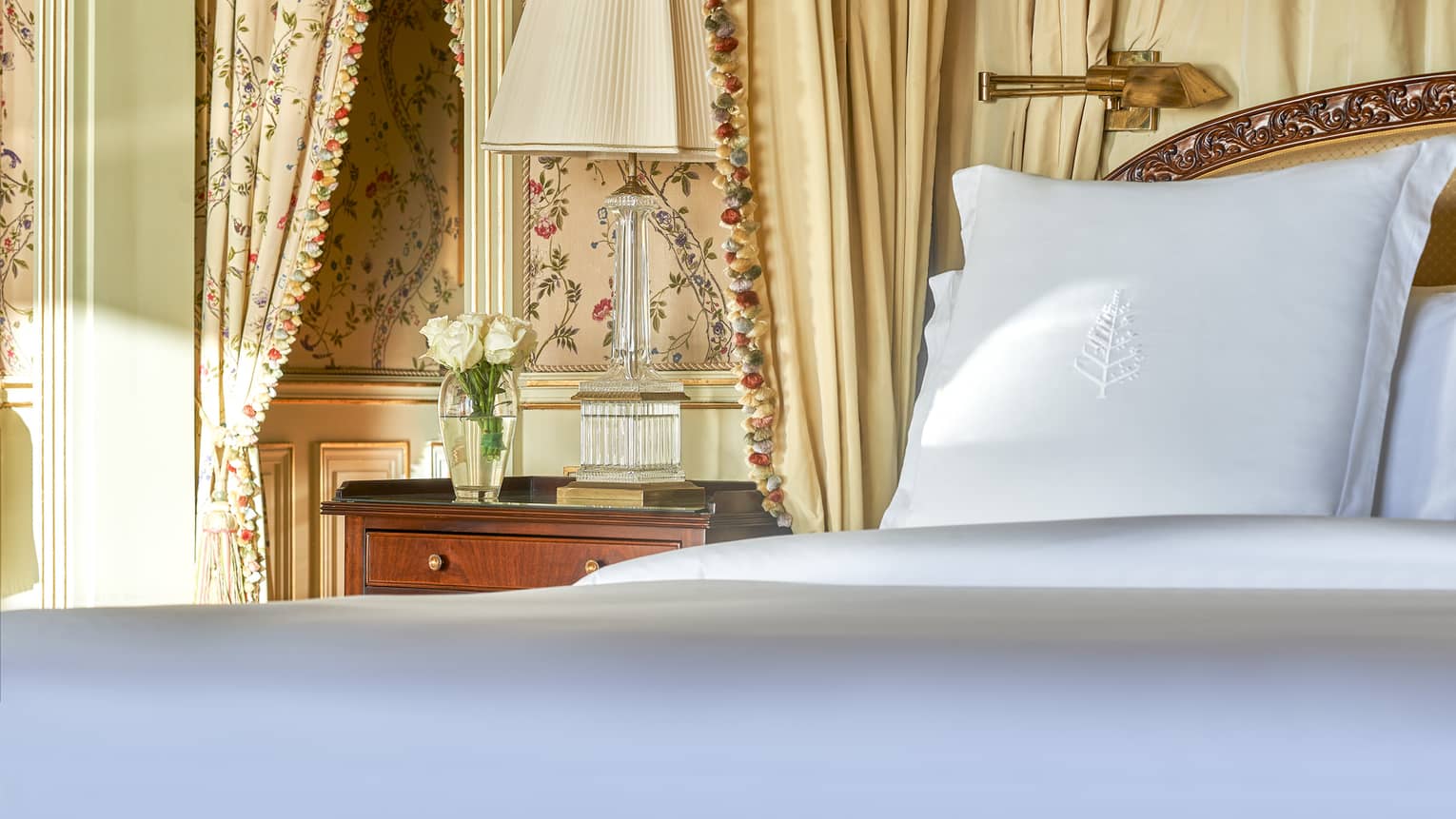 Close-up of bed with white linens, cherry side table, patterned curtains and wallpaper