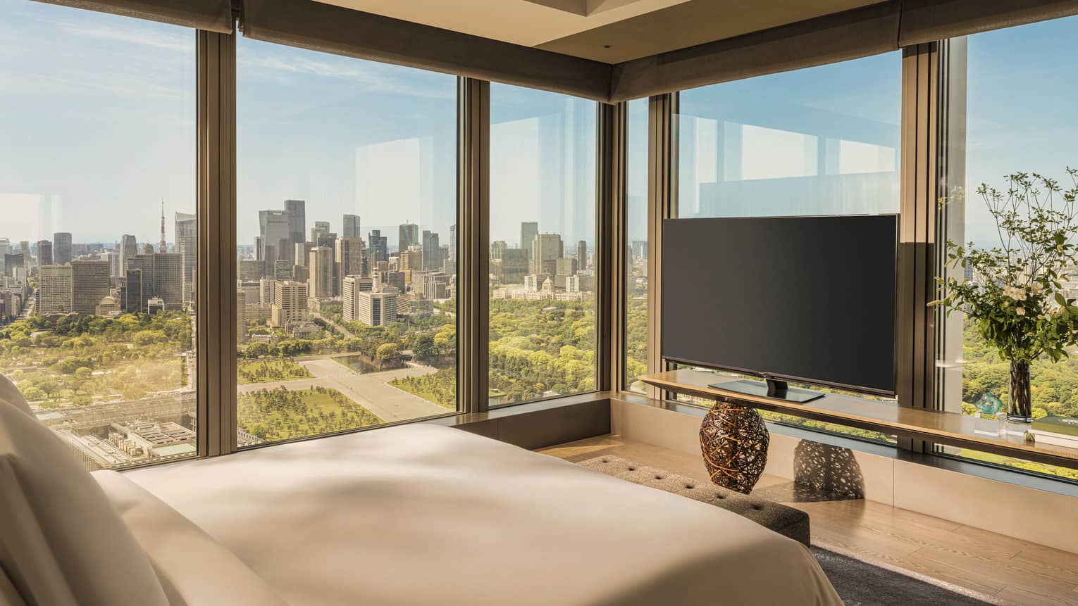 Luxury hotel room featuring king bed, TV and two walls of windows looking out to city and gardens