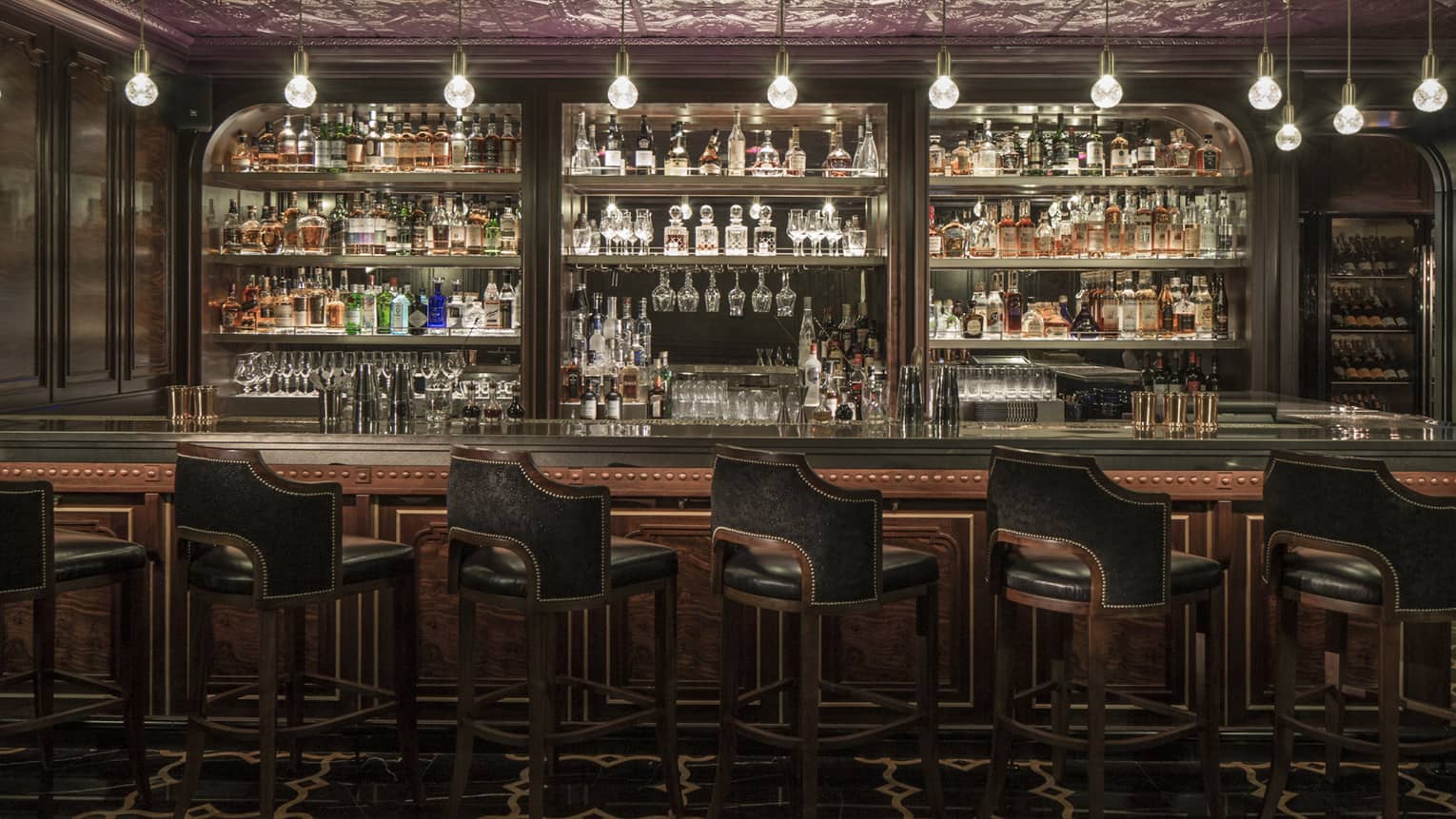 Dimly lit bar counter with black leather bar stools in front and mirrored shelving holding various bottles of spirits behind the bar