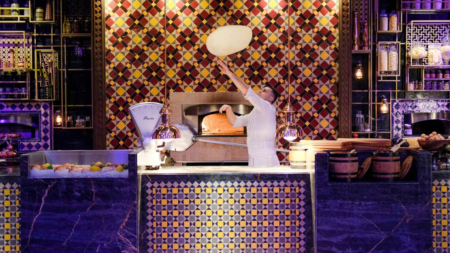 Chef tosses dough above colourful tile counter in front of wood-burning pizza oven