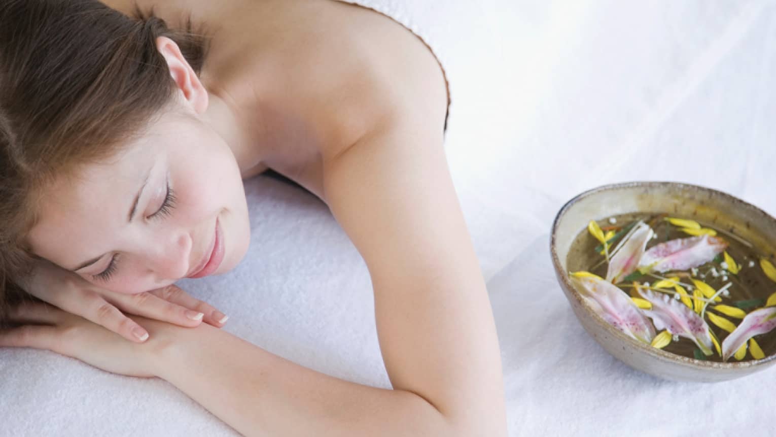 Smiling woman rests on spa table with head on arms beside ceramic bowl with water, flower petals