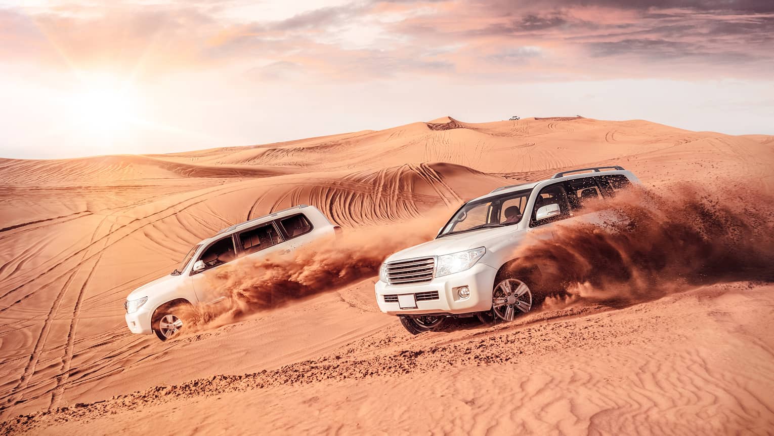 Sand flies from tires of two white SUV's driving across dunes