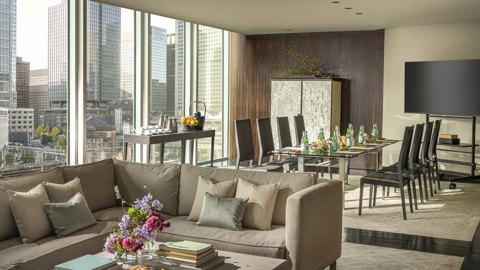 Living area with a large window, city skyline view, beige sectional sofa, table with refreshments, dining area with black chairs and home theatre
