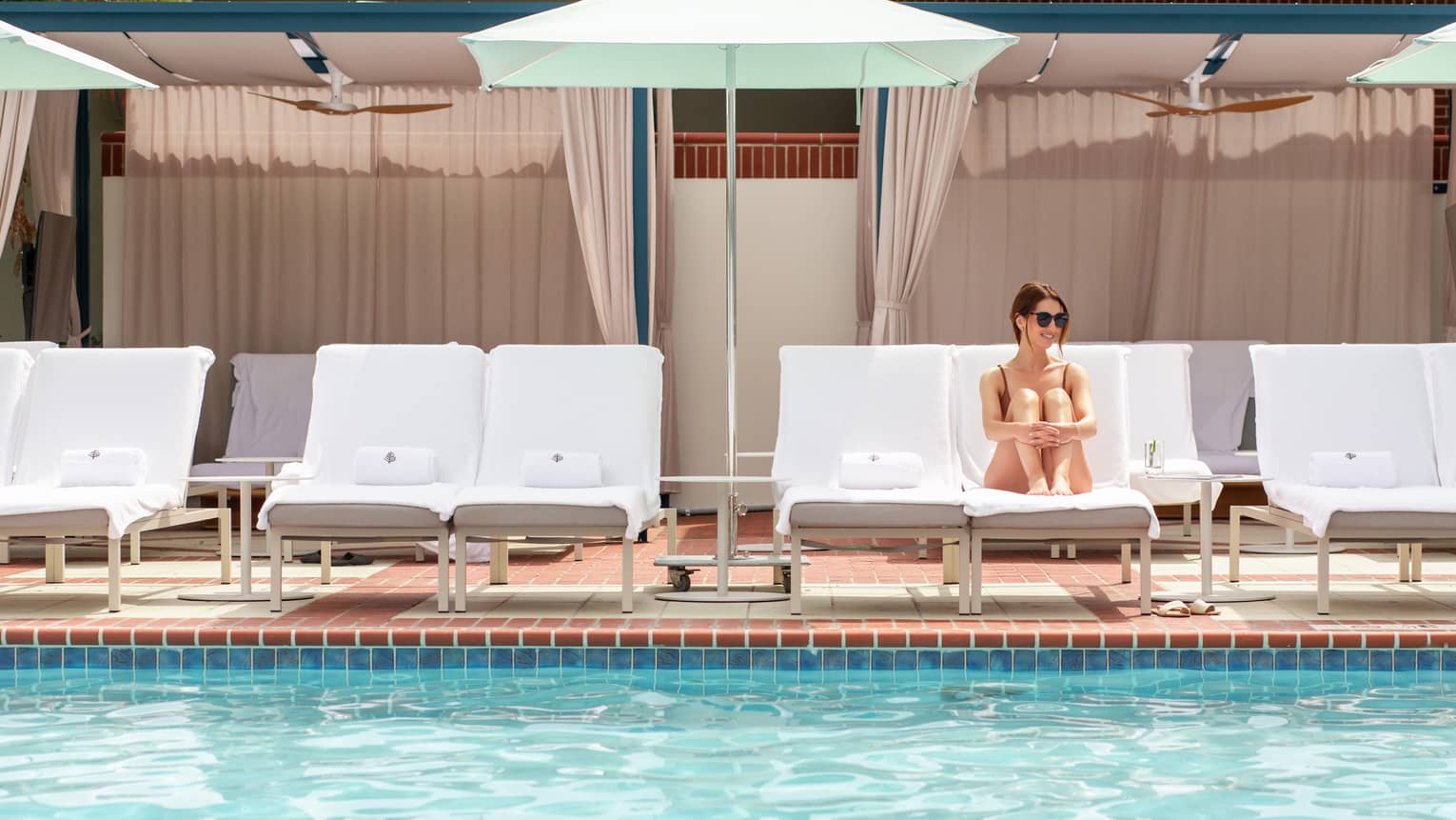 A woman resting on a lounge chair under an umbrella near an outdoor pool.