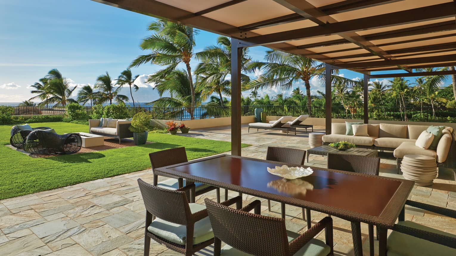 Covered patio with dining table, lounge seating and ocean view surrounded by palm trees and greenery
