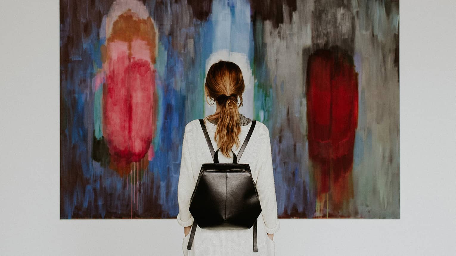 A person with a backpack standing in front of an abstract painting in an art gallery.