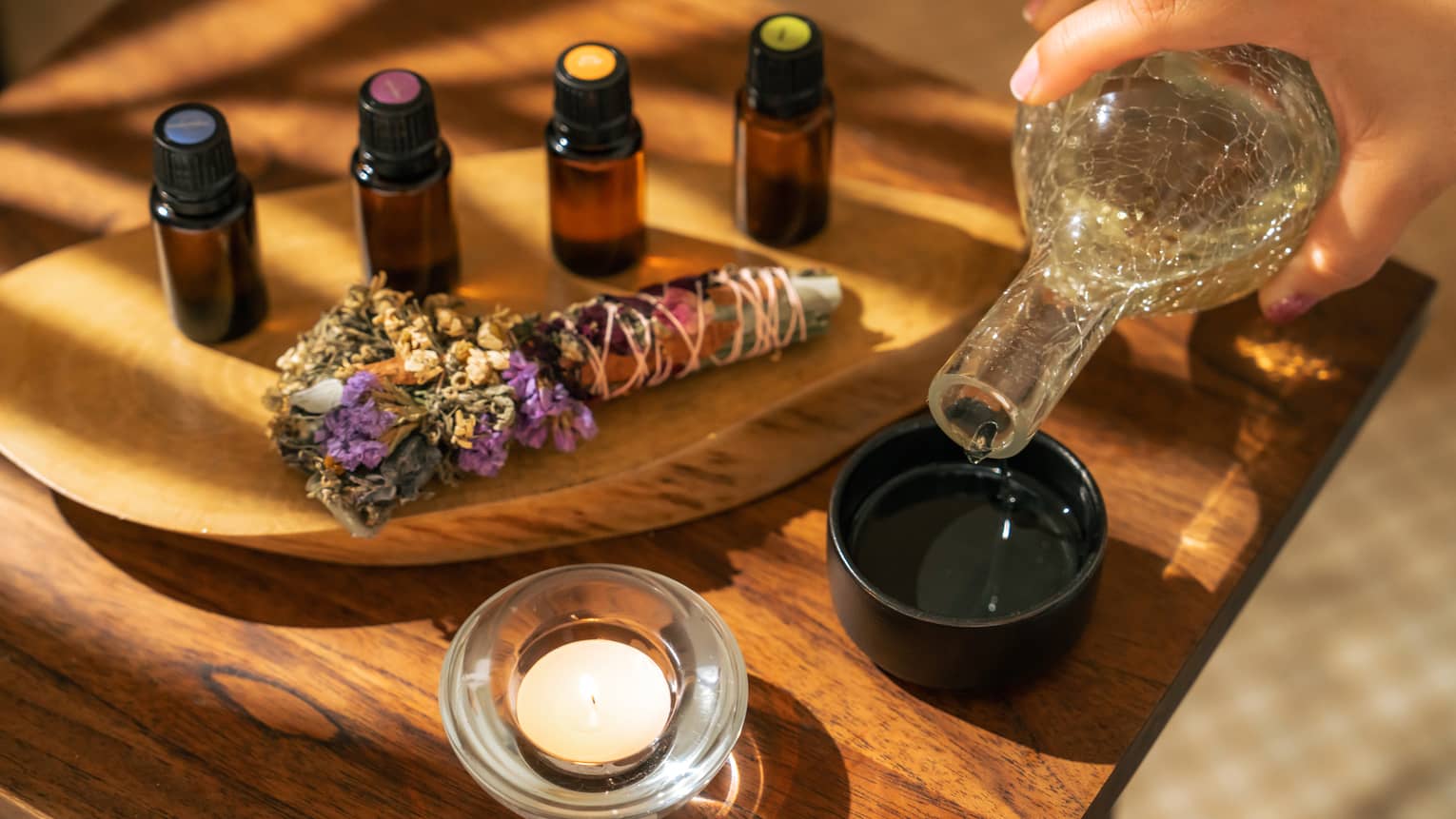 Four small bottles of essential oils and a sage-and-floral bundle stick lie on a wooden platter next to a hand pouring liquid into a small black bowl next to a votive candle