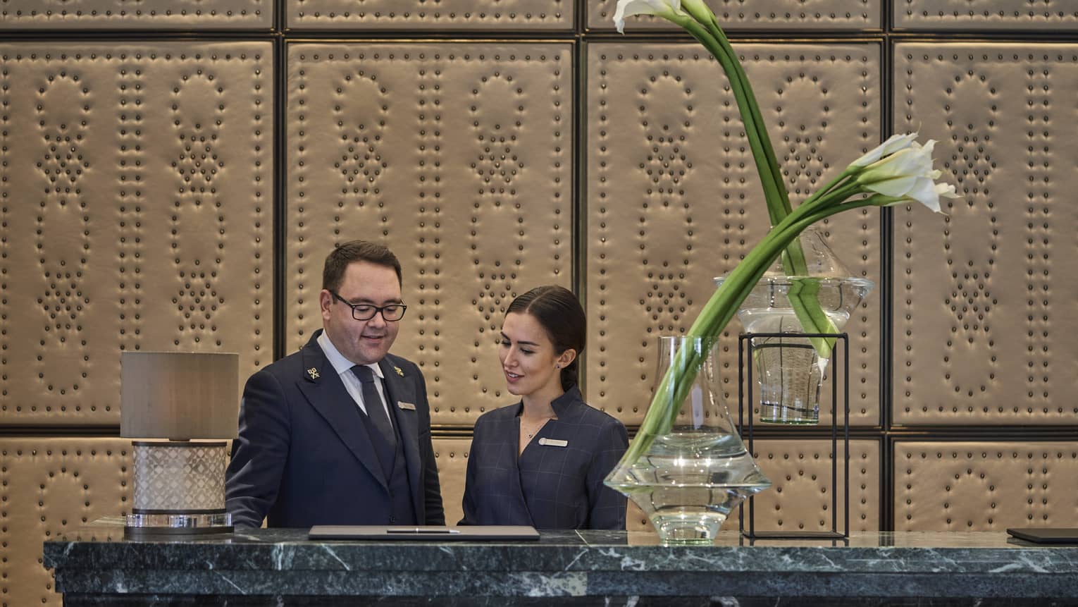 Two people stand behind a reception desk with flowers in a vase in the foreground and textured wall panels in the background