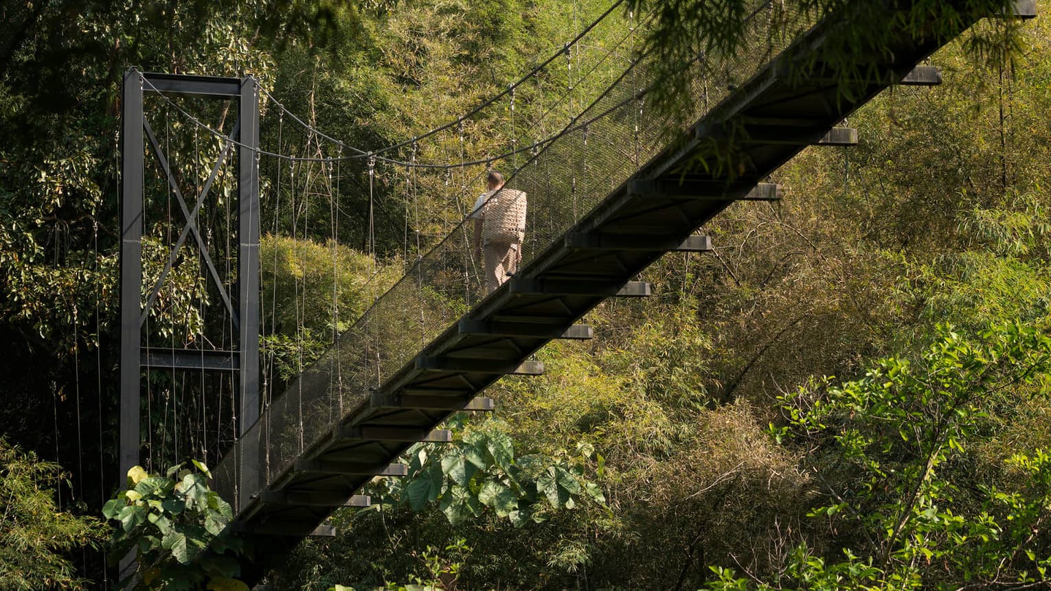Man with woven basket on back walks across suspension bridge over tropical trees